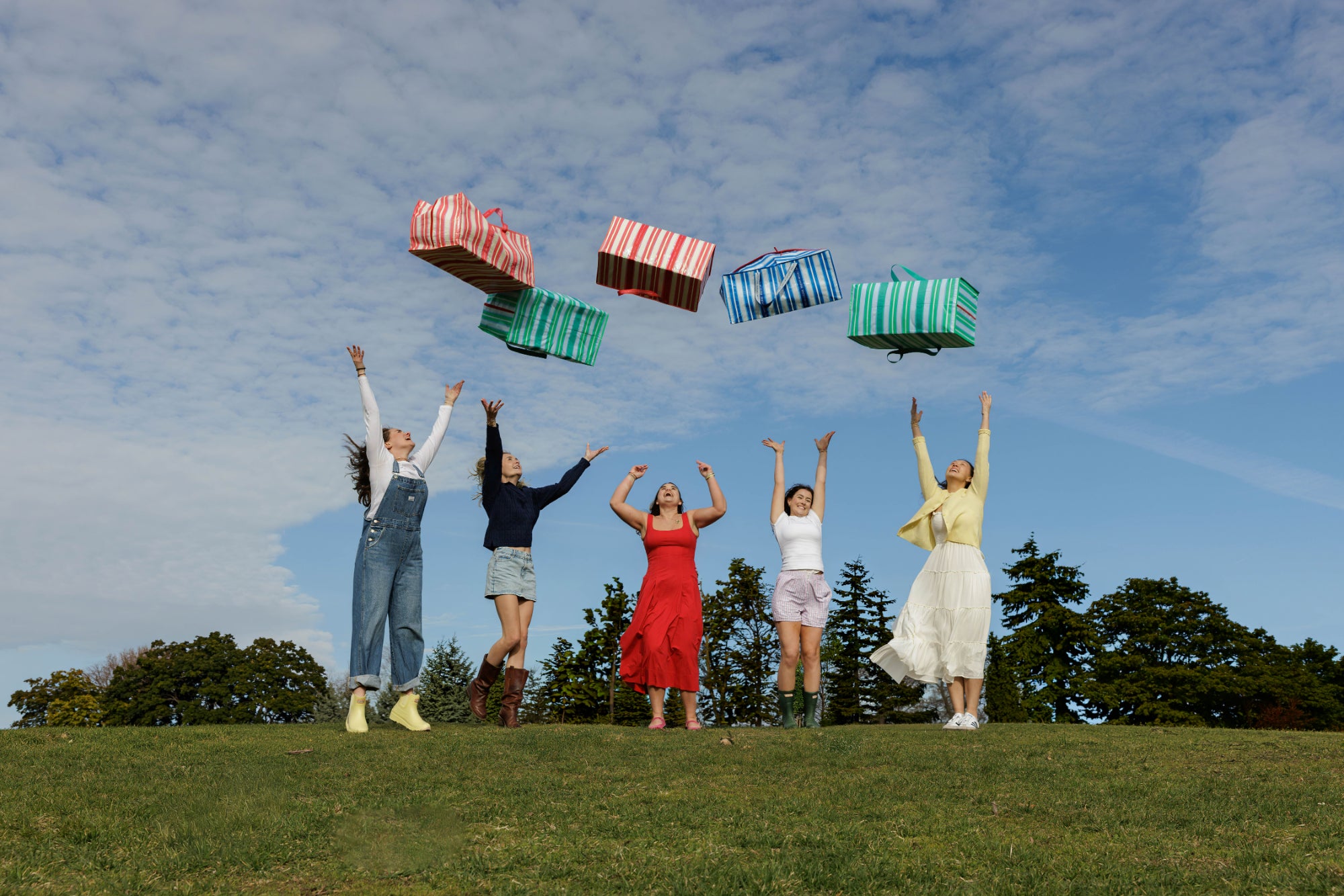 Five women on a grassy hill with colorful and stylish TATUM LOUISE moving bags above them against a blue sky.