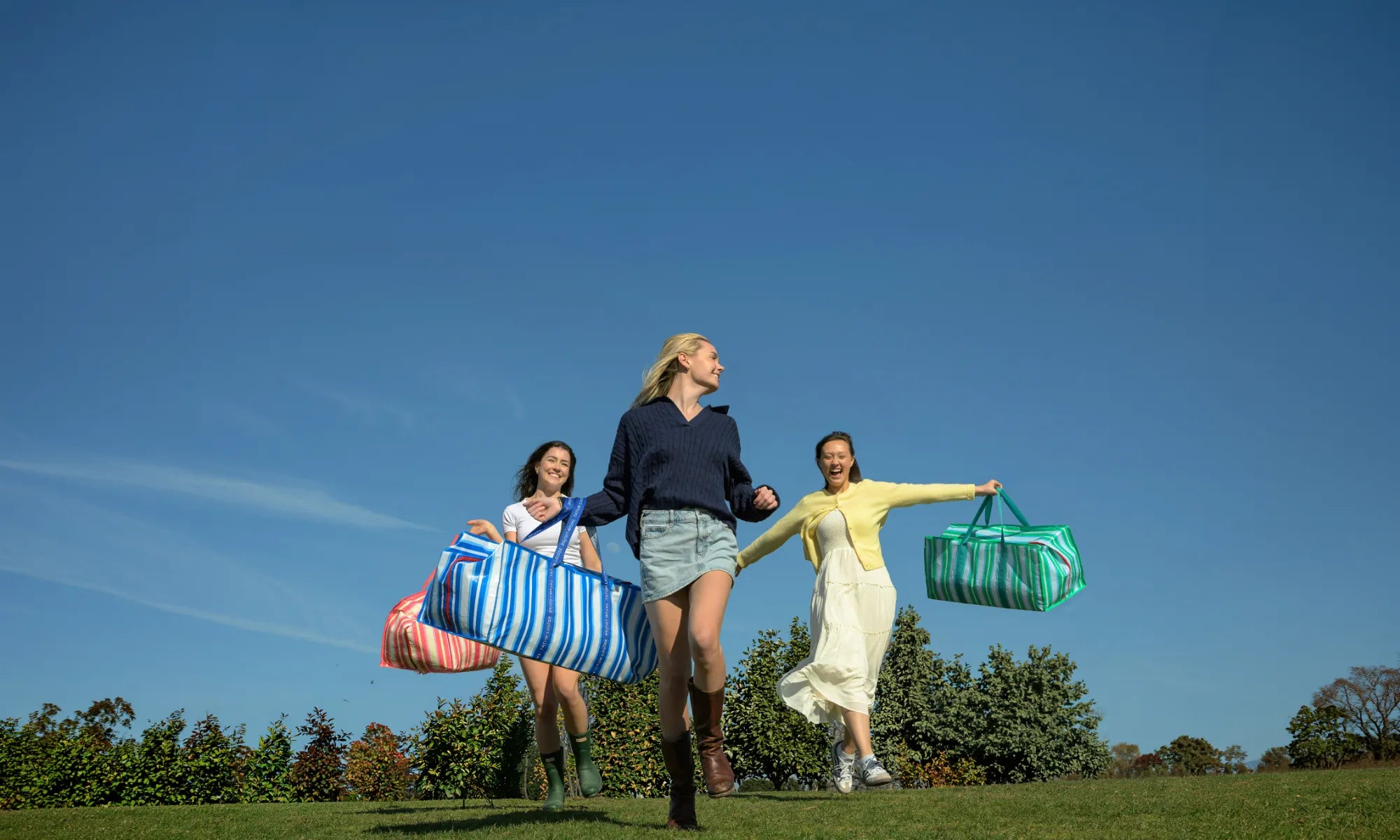 Three women running outdoors with TATUM LOUISE reusable eco-friendly bags against a clear blue sky.